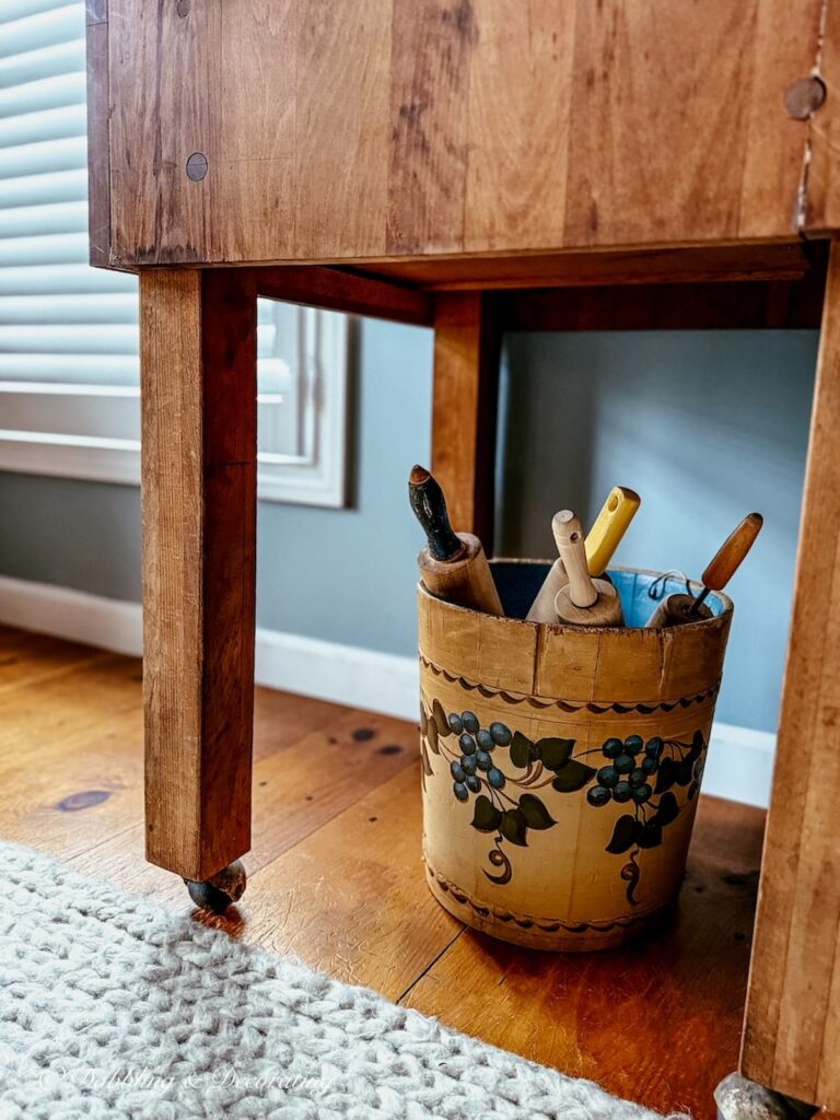 Antique sap bucket filled with vintage rolling pins underneath a wooden butcher block in a kitchen scullery.
