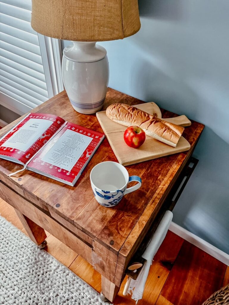 Antique wooden butcher block styled with a thrifted lamp, open cookbook and breadboard with French bread and apple looking down in kitchen scullery.