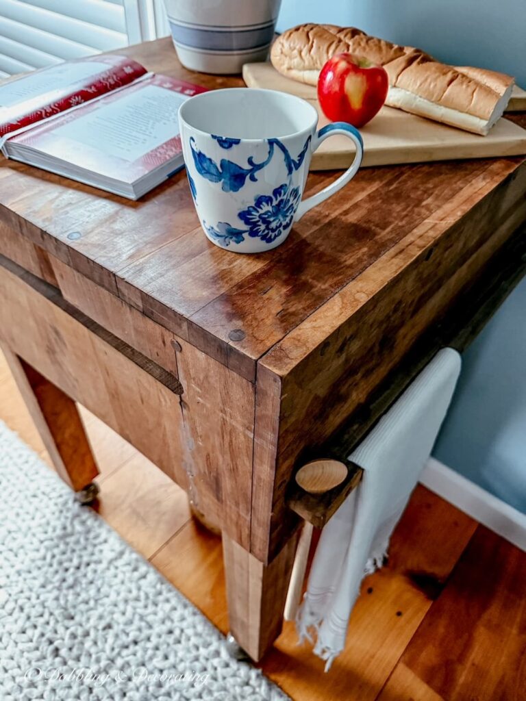 Looking down upon a vintage butcher block styled with a small lamp, breadboard with apple and French bread, open cookbook and coffee mug in scullery.