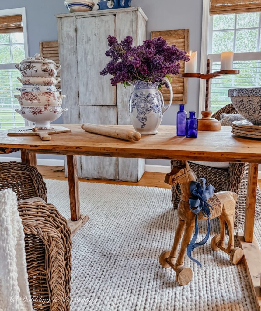 Vintage kitchen-inspired scullery with long wooden table styled with lilacs, a stack of French soup tureens and a large blue cupboard.
