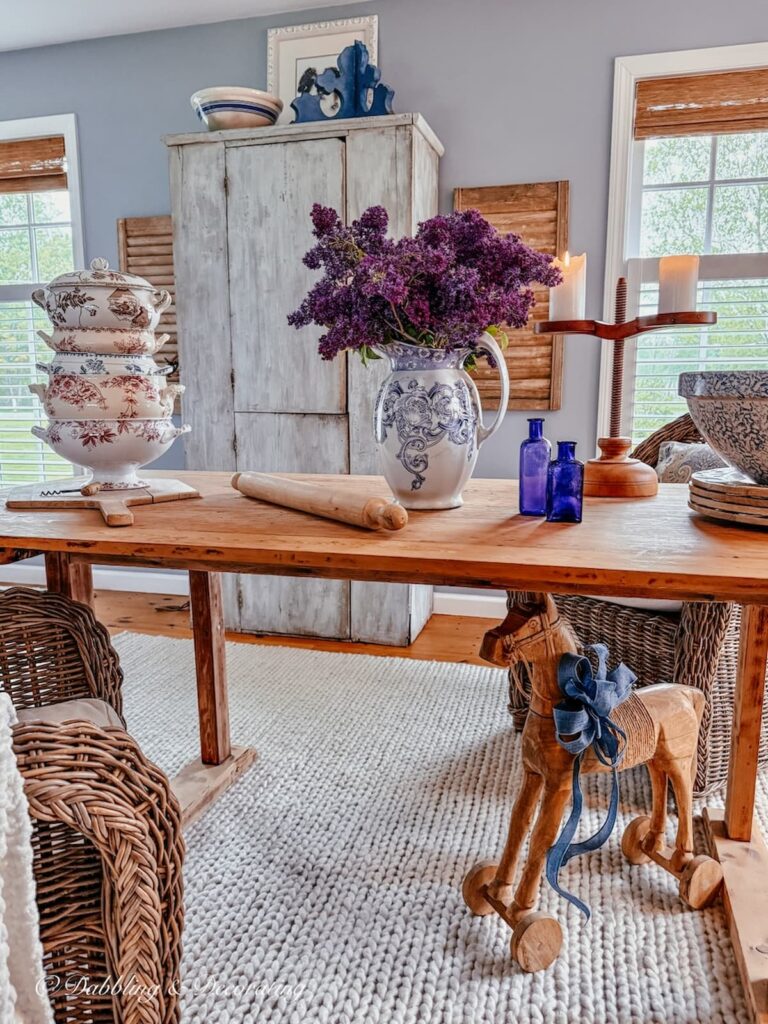 Vintage kitchen-inspired scullery with long wooden table styled with lilacs, a stack of French soup tureens and a large blue cupboard.