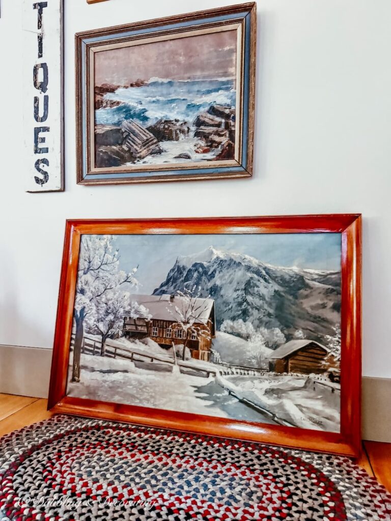 Antique painting in vintage wooden frame of snowy scene and chalet sitting on the floor of our Maine home on a braided rug leaning up against the beginning of a gallery wall.