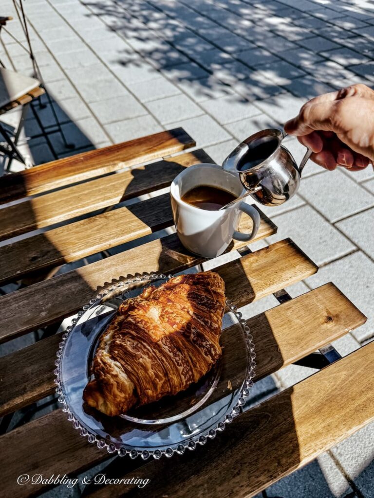 Outdoor cafe table with French croissant, coffee and hand pouring cream in Magog, Quebec.