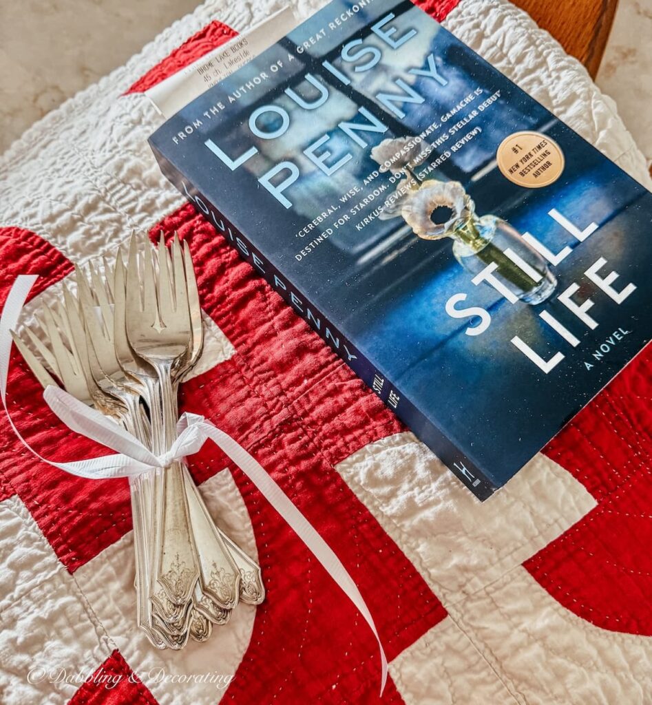 Set of silver forks tied with a ribbon next to Louis Penny's Still Life book on red and white quilt from an antiquing road trip.