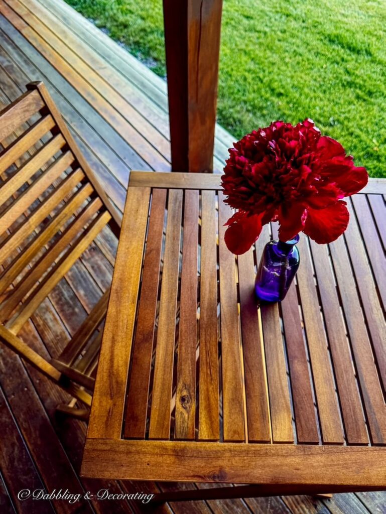 Teak bistro table with single chair and a purple bottle with one red peony centered on covered porch table.