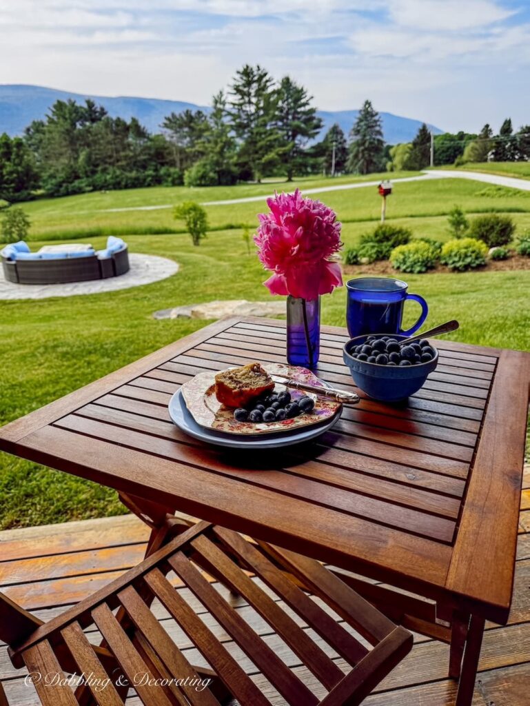 Teak bistro table for one set for breakfast with thrifted finds on our covered porch in Vermont with mountain views.