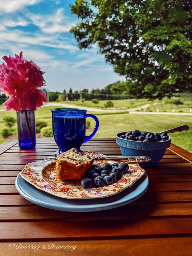 Table for one with thrifted plates, mug, bowl and bottle with peony set for breakfast on covered porch in Vermont.