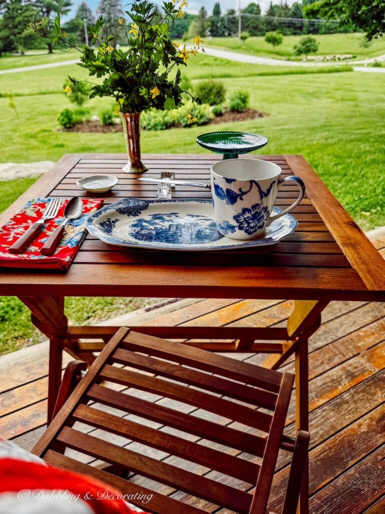 A teak bistro table with chair set for breakfast with blue and white thrifted dinnerware on covered porch for summer in the mountains.