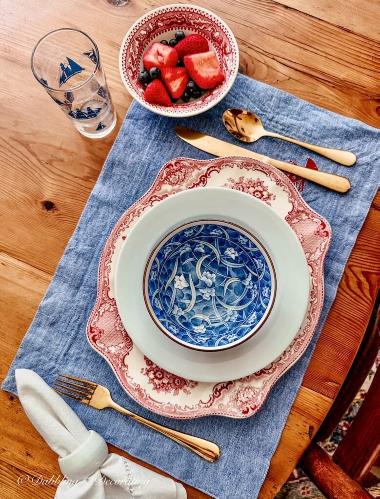 Vintage patriotic Place Settings with layered dishes on a denim placemat with gold flatware and sailboat juice glass.