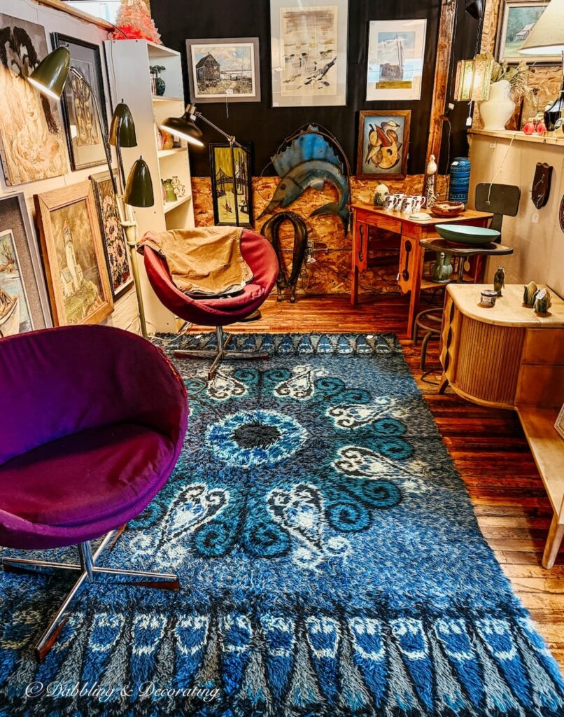 Mid century modern booth display with vibrant blue rug, two purple vintage urban chairs and colorful wall paintings at Portland Antique Mall in Maine.