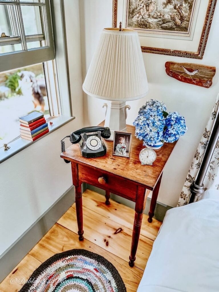 Antique bedroom wooden side table decorated with blue hydrangeas, old telephone and open window and braided rug on floor for vintage summer decor ideas.