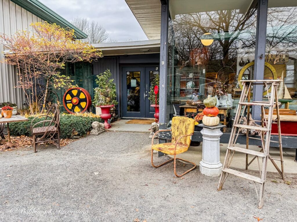 Fall vintage booth displays outside an antique store in the Berskhires of Massachusetts with old ladder, white pedestal and stacked pumpkins with rustic wrought iron yellow chair.