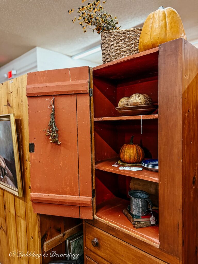 burnt orange hutch with door open and shelves styled with pumpkins, baskets and antiques. Simple and classic fall vintage booth styling.