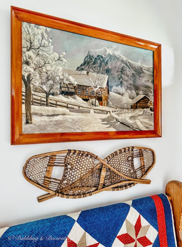 Antique painting of snow scene with cabin hanging on entryway wall with church pew, snowshoes mounted on wall, red, white, and blue vintage quilt