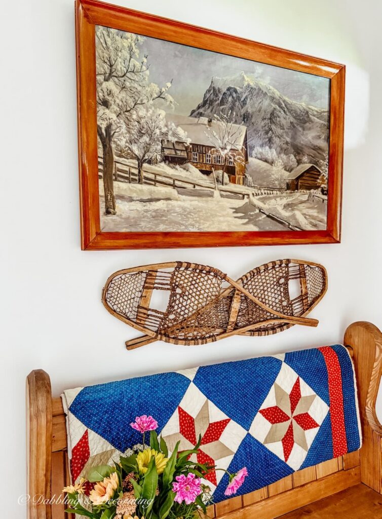 Antique painting of snow scene with cabin hanging on entryway wall with church pew, snowshoes mounted on wall, red, white, and blue vintage quilt and summer flower bouquet.