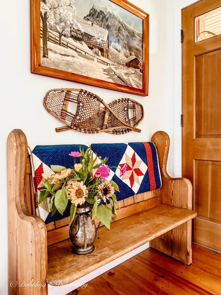 Antique church pew in home's entryway with red, white, and blue quilt, summer flower bouquet and snowshoes criss crossed on wall underneath antique painting with snow scene at home in Vermont.