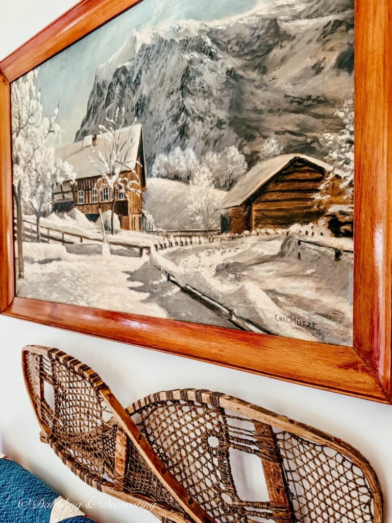 Antique painting with snowy cabin scene in the mountains on wall with criss crossed snowshoes.