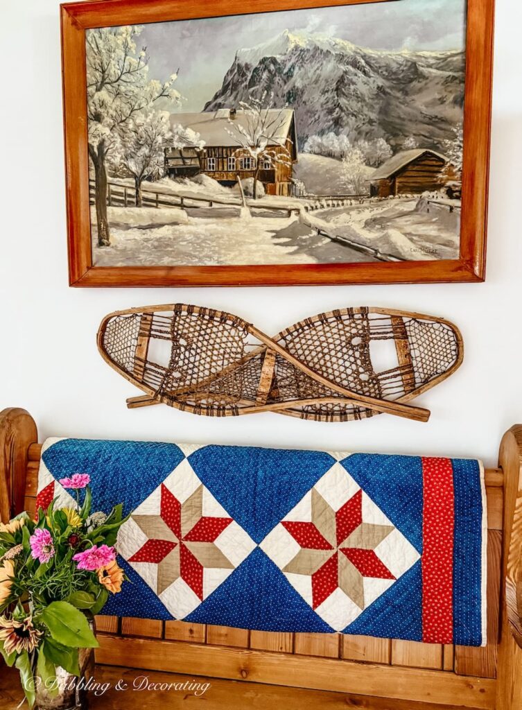 Antique painting of snow scene with cabin hanging on entryway wall with church pew, snowshoes mounted on wall, red, white, and blue vintage quilt and summer flower bouquet.