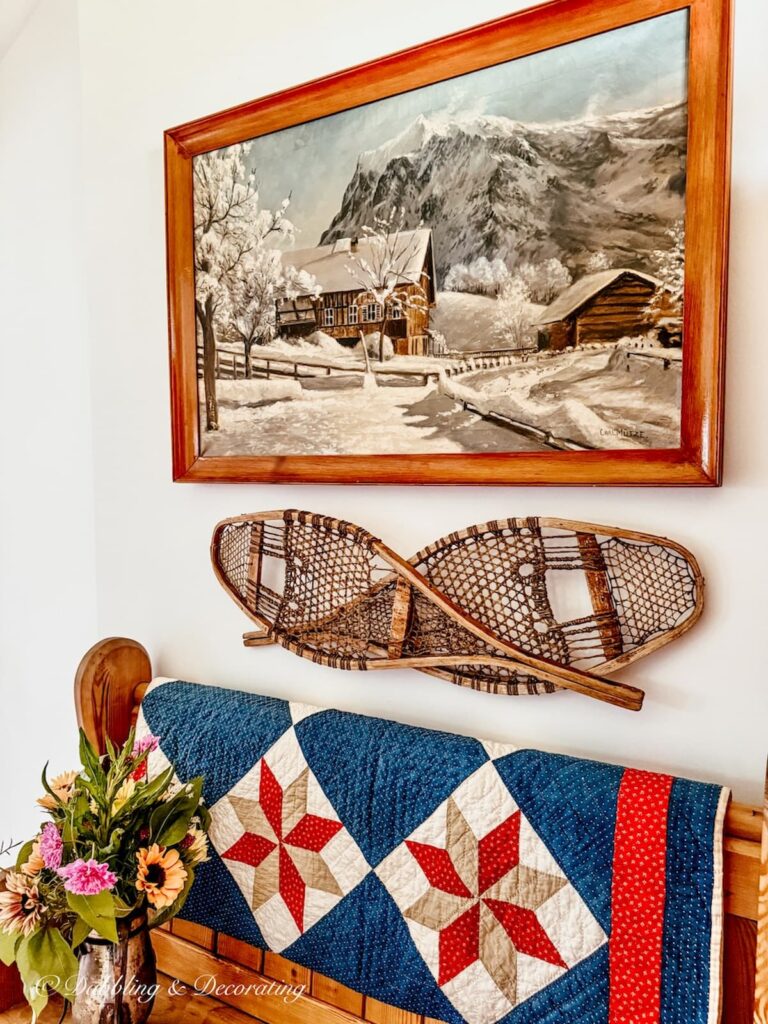 Antique painting with snowy cabin scene on entryway wall with criss crossed snowshoes and church pew with red, white, and blue quilt and summer flowers.