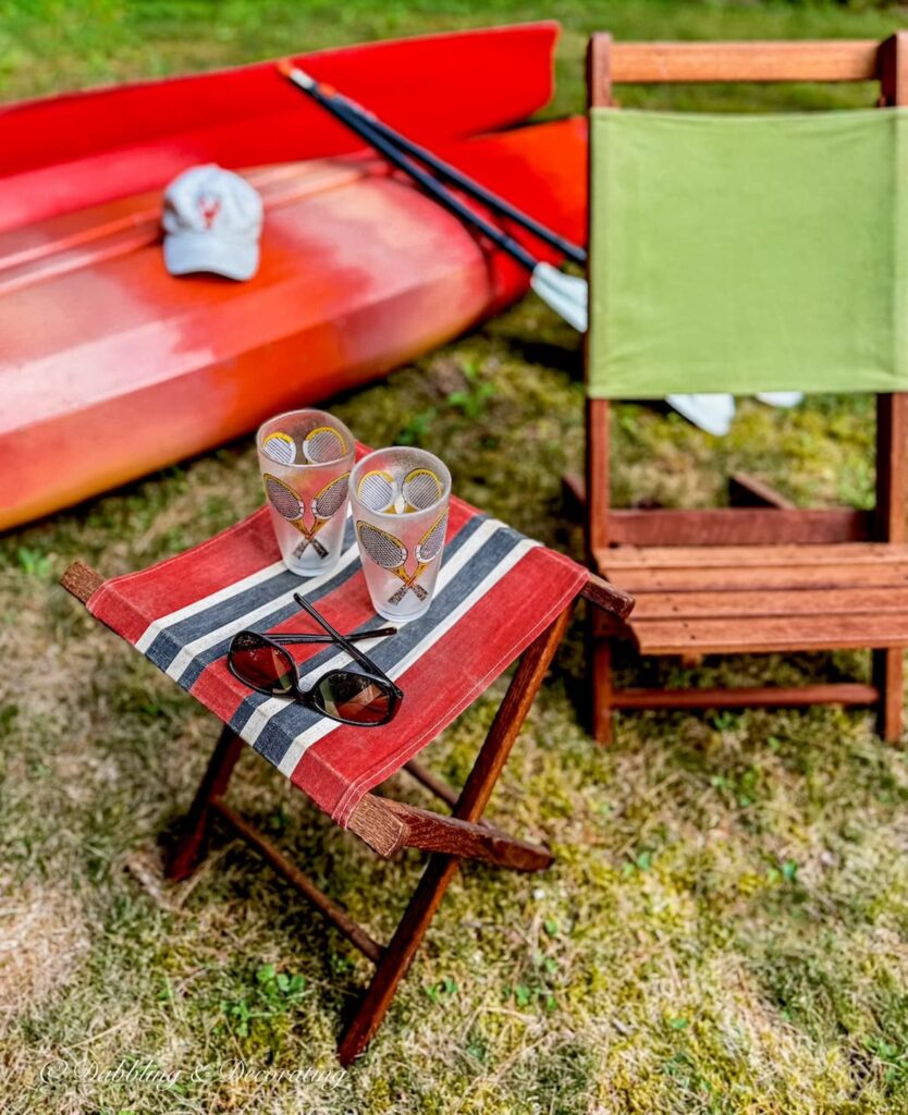 Wooden folding camp chair with green canvas next to a camp stool with vintage glassware and kayaks on a lawn for summer vintage decor ideas.