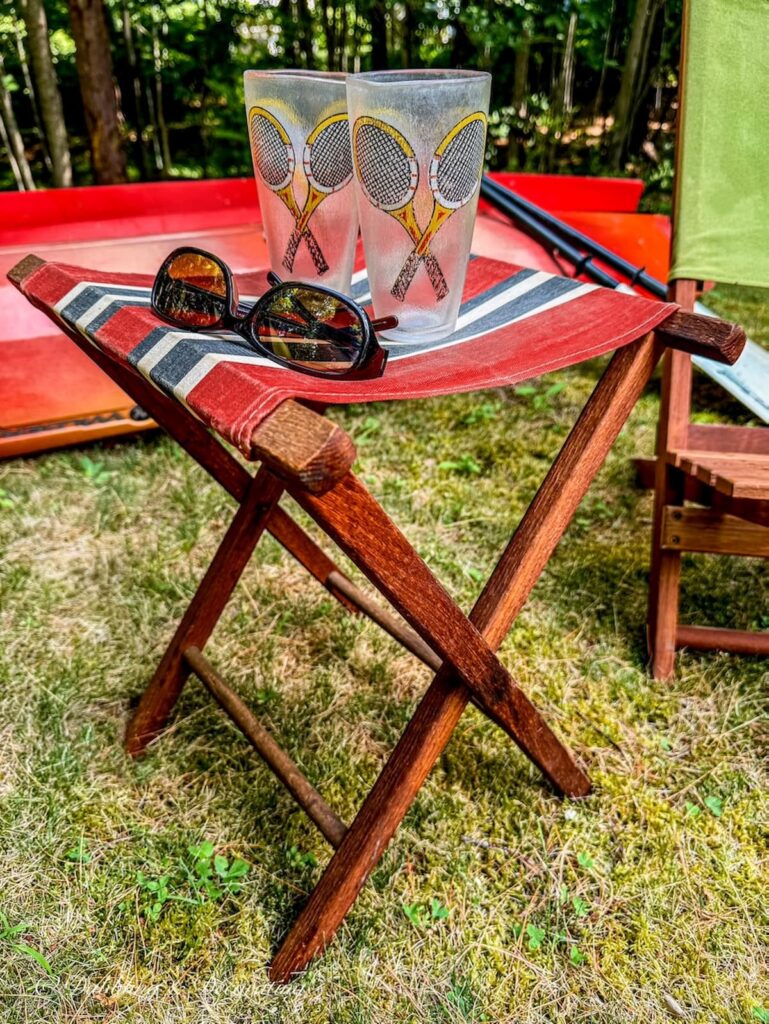 Little red canvas retro camping stool with old tennis plastic glasses and sunglasses in front of outdoor kayaks on the lawn.