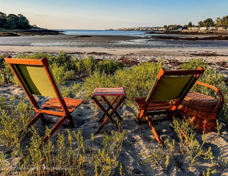 Two retro wooden camp chairs on the beach with a small camp stool and vintage picnic basket at sunset on a beach in Maine with a view.