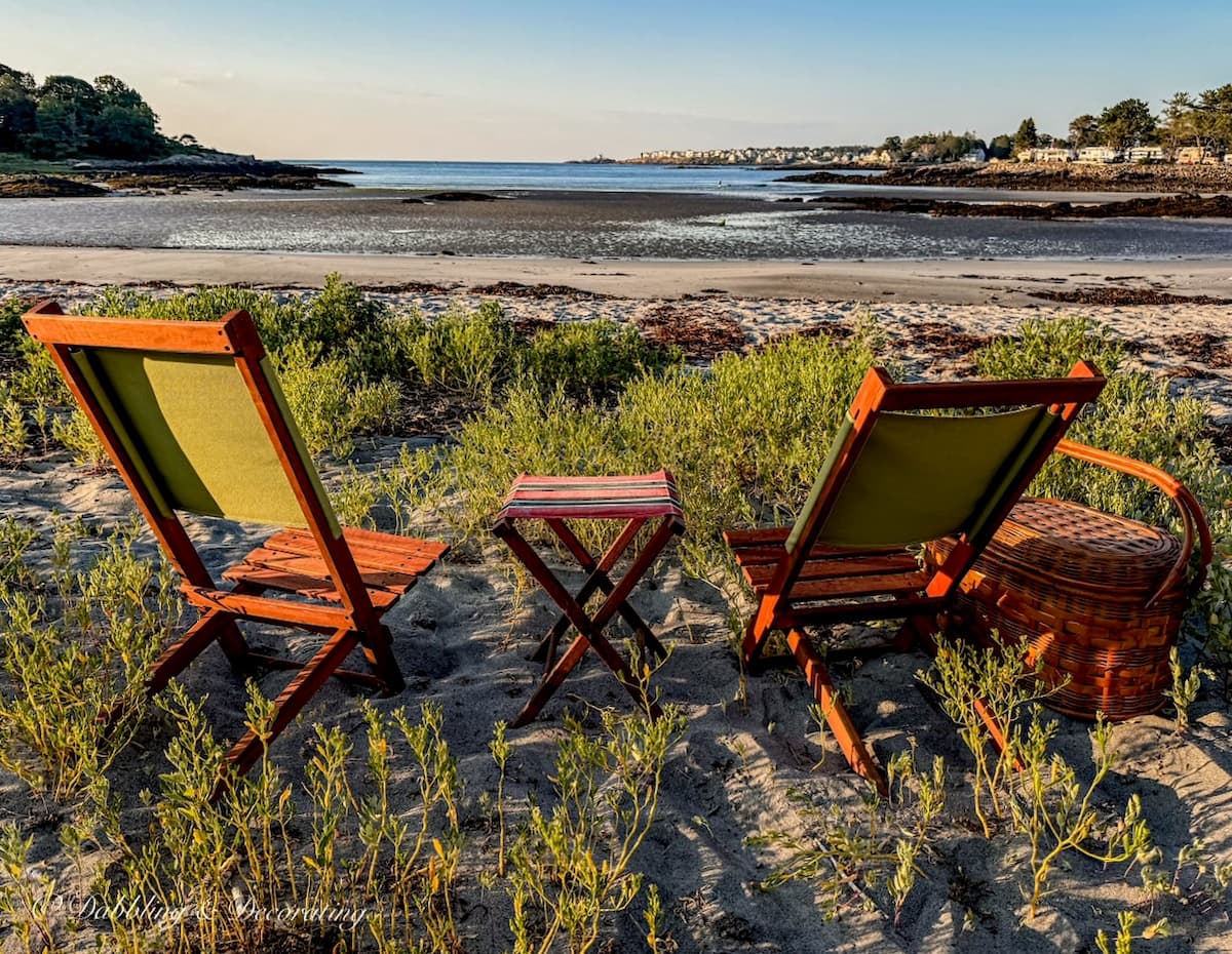 Two retro wooden camp chairs on the beach with a small camp stool and vintage picnic basket at sunset on a beach in Maine with a view.