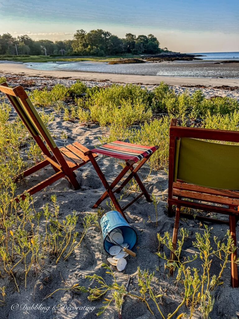 Two retro wooden camp chairs with a small camp stool and blue bucket spilled over with sand dollars on a beach in Maine at sunset.
