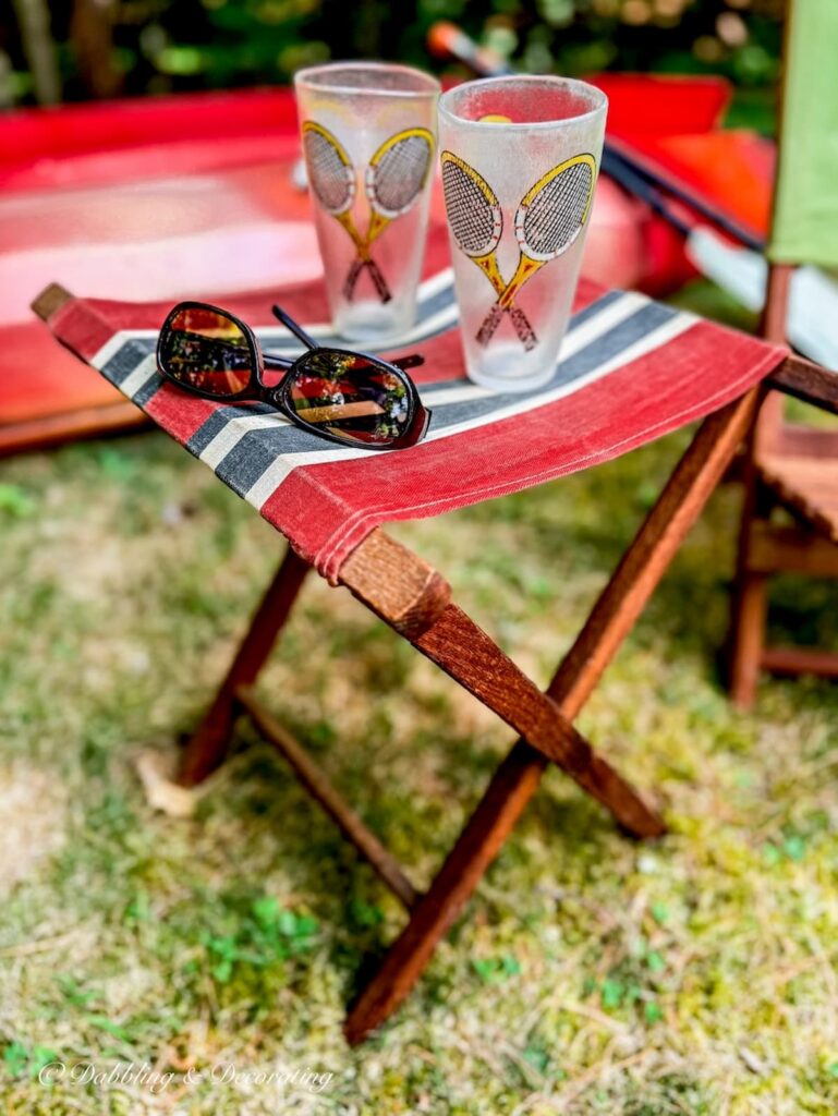 Small vintage wooden camp stand with red, white, and black canvas styled with retro tennis glasses, pair of sunglasses with kayaks in background.