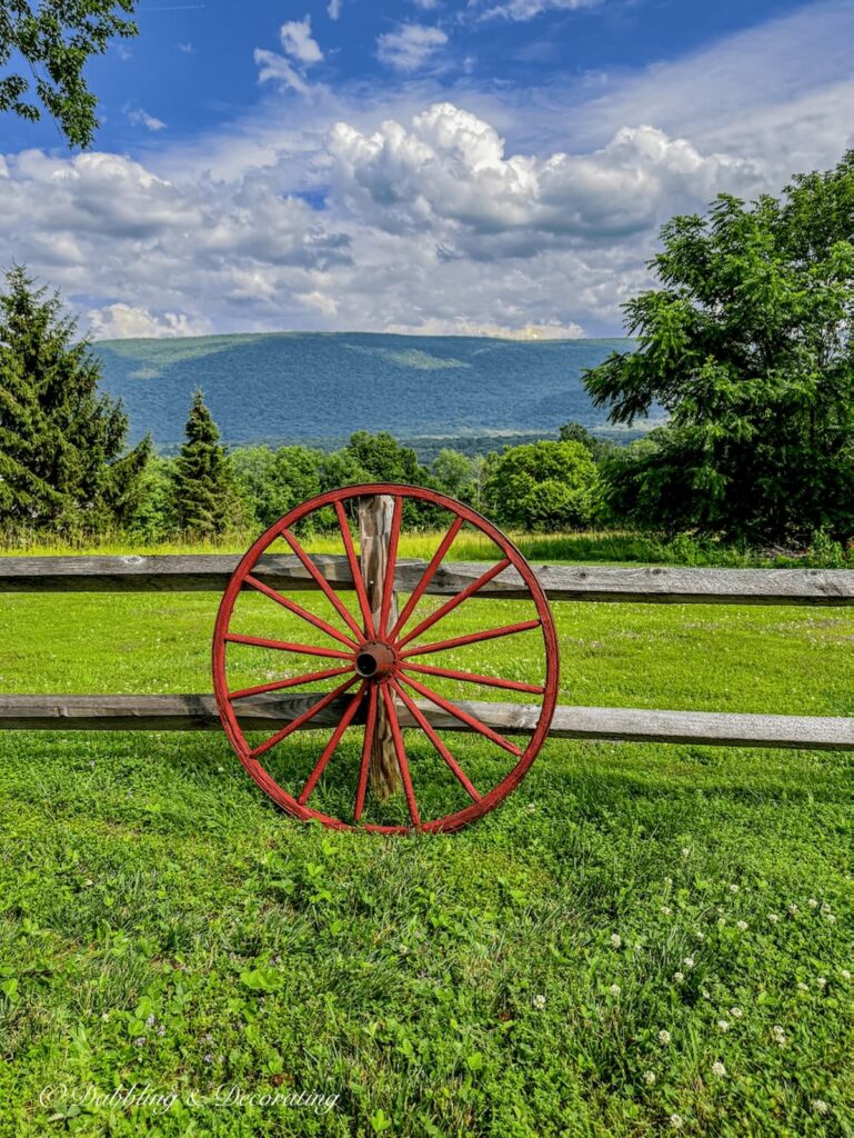Old wagon wheel painted red leaning up against a weathered split rail fence with mountain landscape at Vermont home.