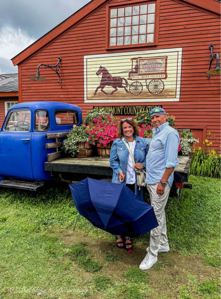 Steve and Ann in front of old blue truck at The Vermont Country Store with flowers and umbrella on a rainy day.