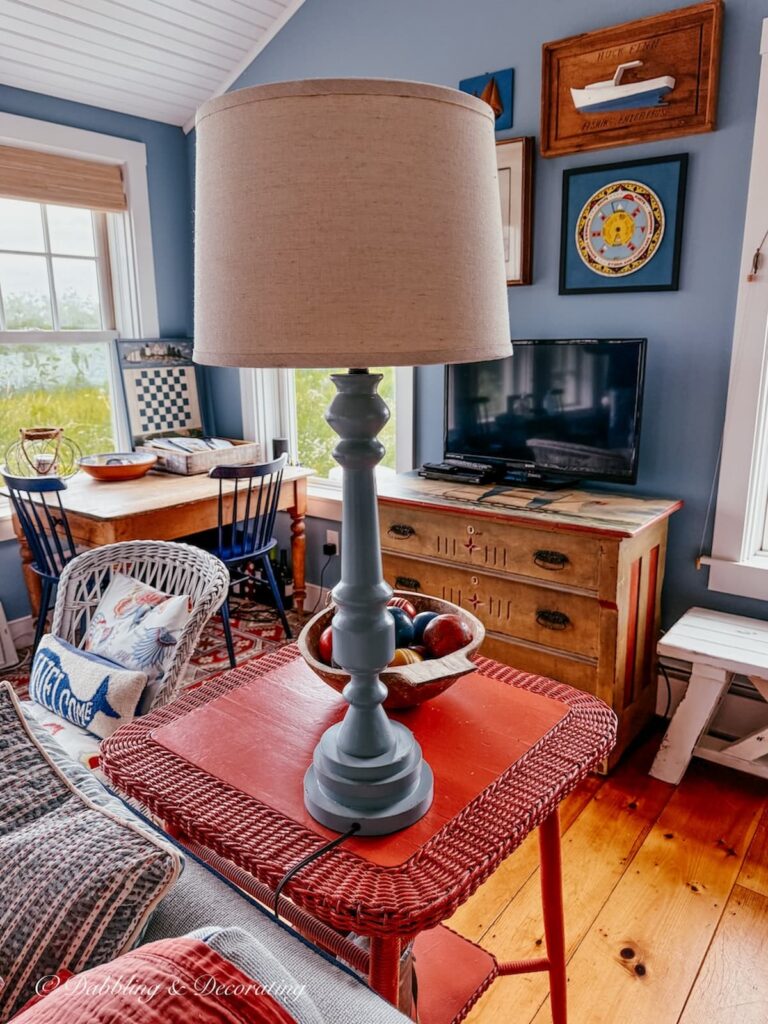 Salmon colored wicker side table with blue lamp in colorful guest cottage cottage with antiques and gallery wall.