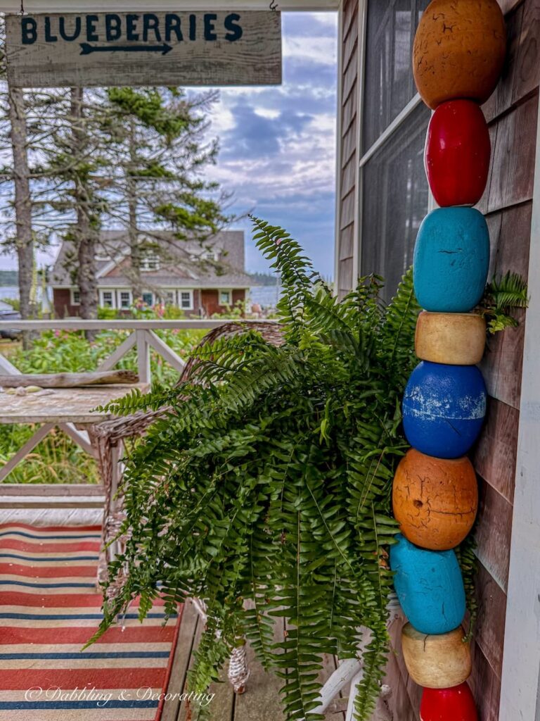 Colorful ocean buoys hanging from a rope on a guest cottage porch with a fern and a view of the ocean and property home with a vintage Blueberry sign.