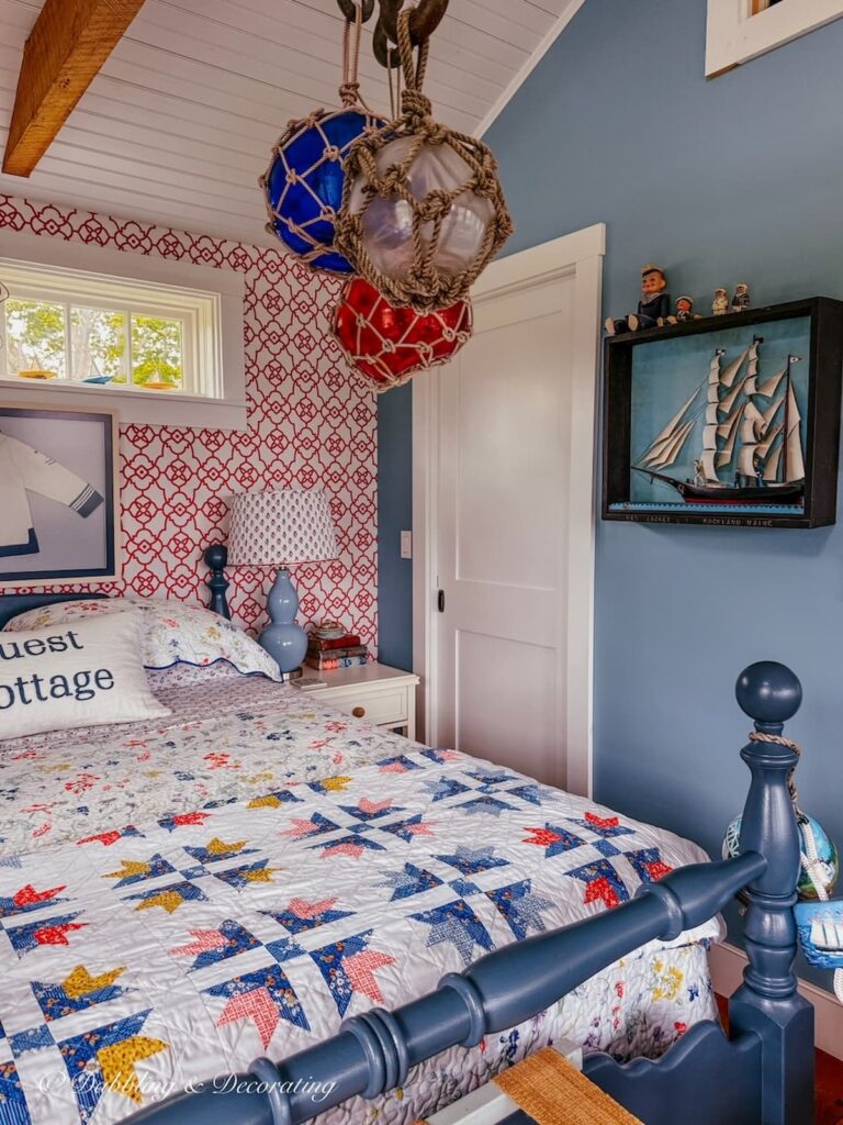 Guest cottage bedroom with queen blue bed layered with quilts and three vintage glass fishing floats wrapped in rope netting hanging from ceiling beam on old anchors.