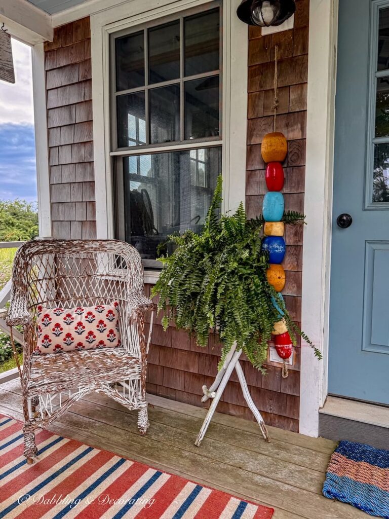 The outdoor landing of a Maine guest cottage styled with colorful details like a wicker chair with cushion, red striped rug and hanging colorful lobster buoys with summer fern and a blue front door.