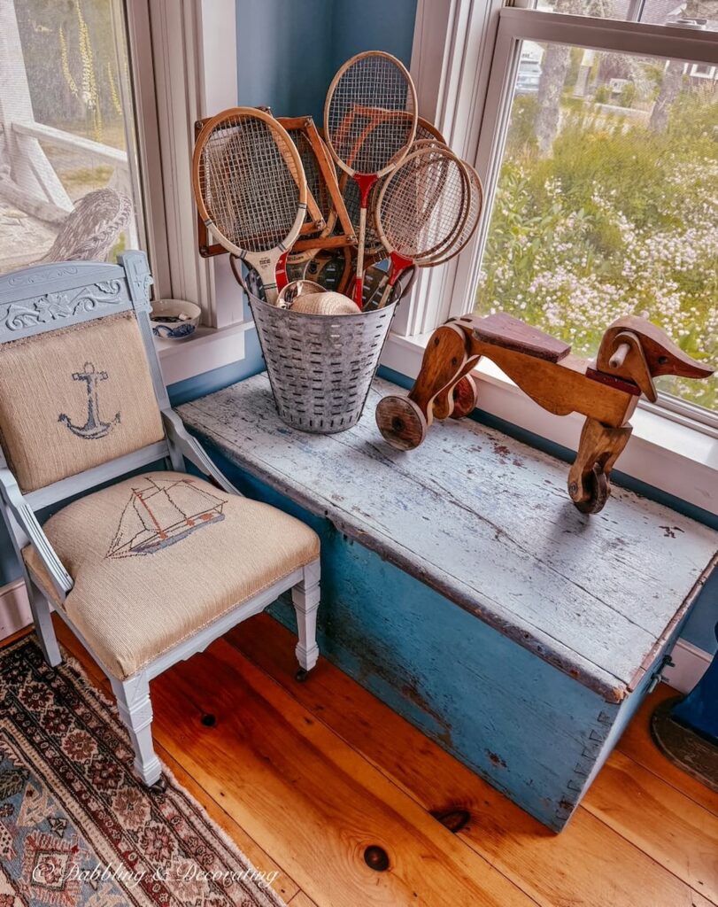 Vintage tin filled with tennis rackets and badminton rackets on blue trunk with wooden dog toy and coastal blue chair in corner of guest cottage.