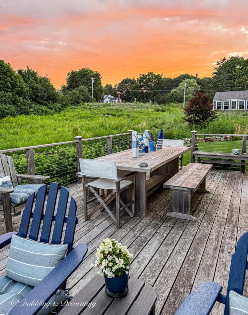 Sunset at a home in coastal Maine during the summer on porch with blue Adirondack chairs and large picnic table and guest cottage off in the background.