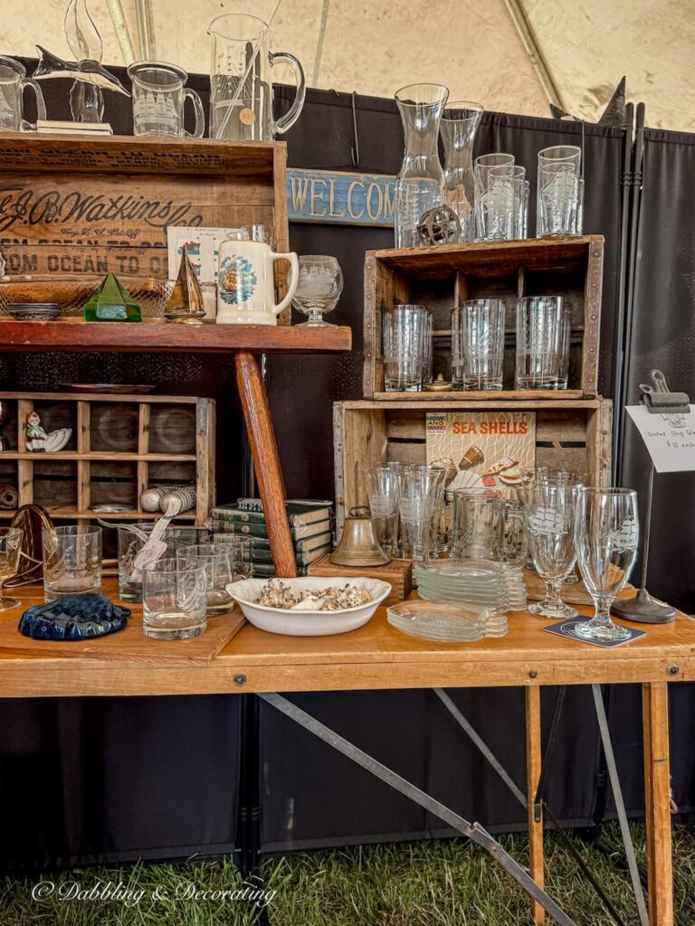 Antique booth display on old wallpaper table showcasing vintage glassware and crystal in contrast with wooden crate displays.