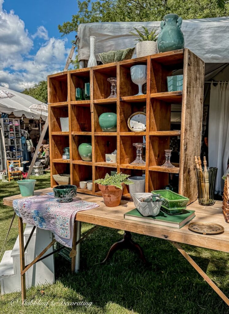 Large wooden vintage crate with cubby holes filled with vintage pitchers, mugs, bowls, and more on vintage wallpaper table in antique booth display at Vintage Bazaar.