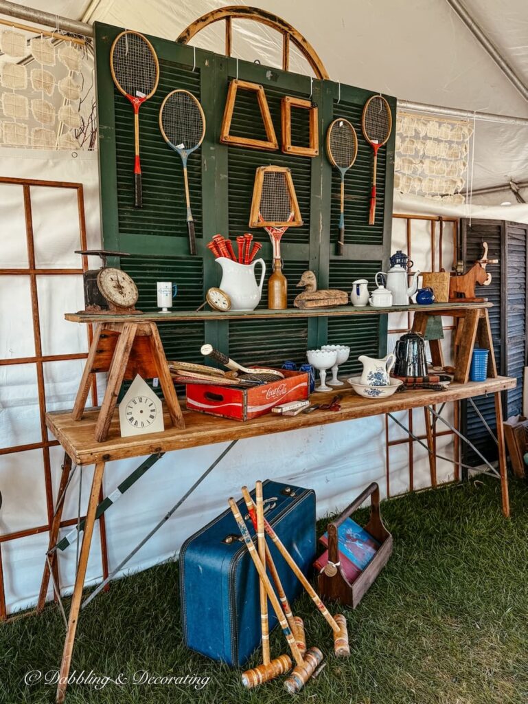Creatively styled tennis rackets hung on a backdrop of old green shutters on top of vintage wallpaper table with old suitcase, crochet mallets and more at an outdoor summer market.