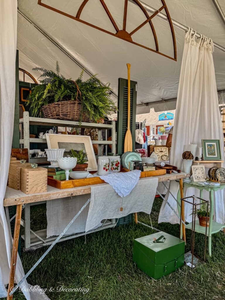Antique booth display outdoors with vintage wallpaper table display architectural storage hung above and white curtains blowing in the breeze from tent rails.