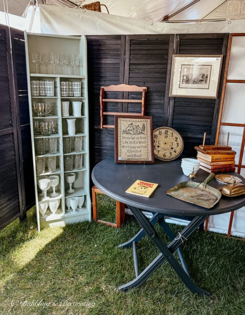 Vintage Bazaar antique booth display with old shutters as backdrop with round dining table, and glassware display.