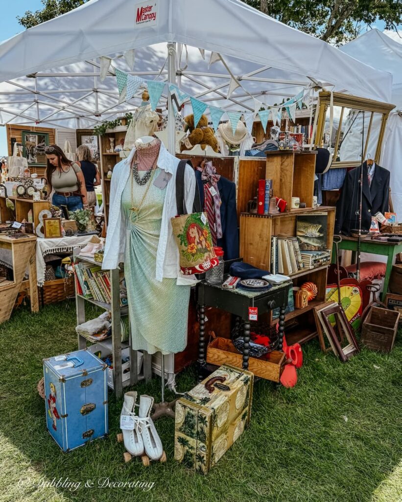 Antique booth display at Vintage Bazaar New England in Mystic, CT with mannequin, old suitcases, roller skates, vintage clothing and more under outdoor tent.