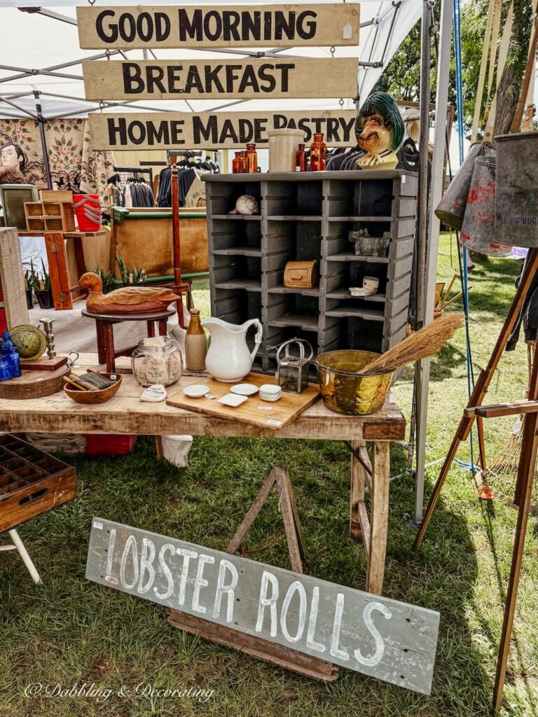 Antique booth display with vintage sign "Lobster Rolls" and other seaside signs and layered vintage displays at outdoor flea market.