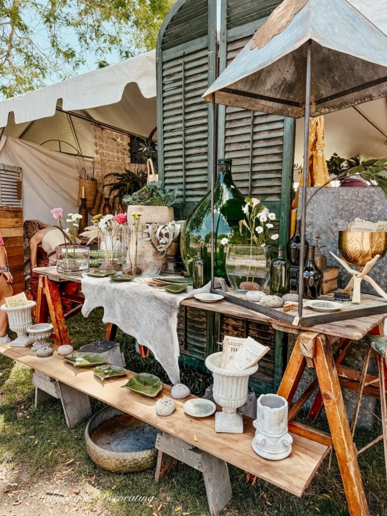 Vintage shutters, vintage wallpaper table displaying vintage dishes, bottles, lamps, linens and more at Vintage Bazaar New England outdoors under tent.