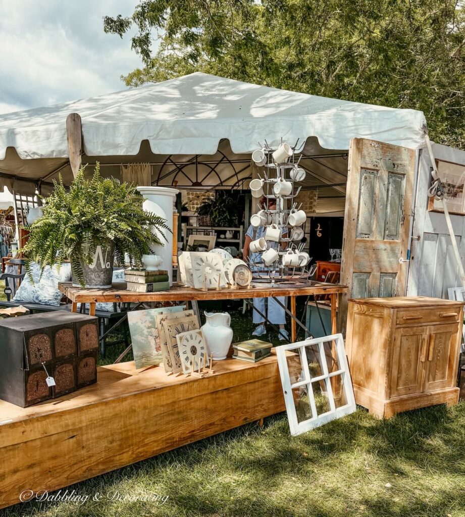 Antique booth displays outdoors at Vintage Bazaar with layered tables, crates, salvaged doors and windows, ferns and more.