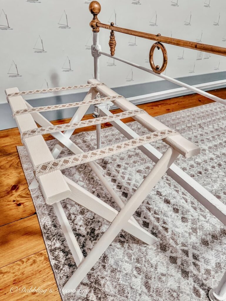 Cream with ribbon DIY folding luggage rack at foot of antique wrought iron white bed in bedroom makeover.