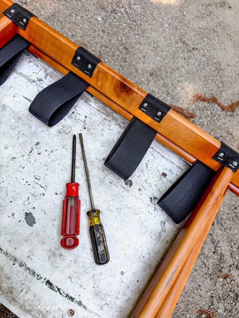 Folding luggage rack on table with visual of screws and staples of webbing to be removed.
