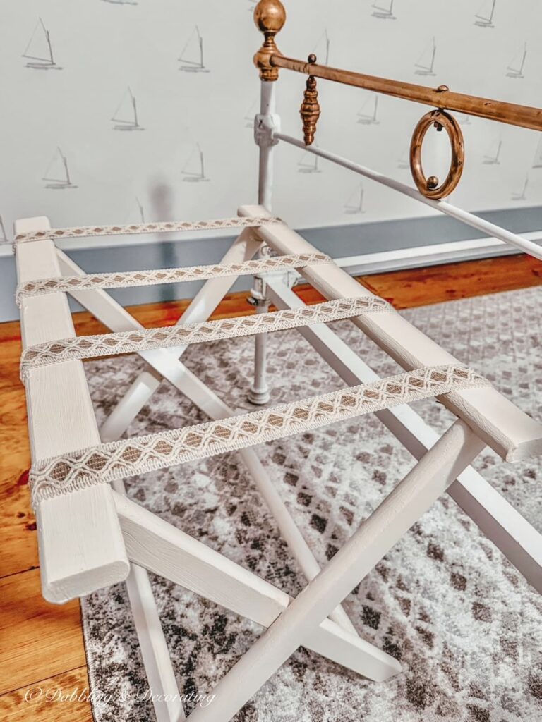 DIY folding luggage rack makeover in cream with ribbon at the end of an antique white wrought iron bed with brass.