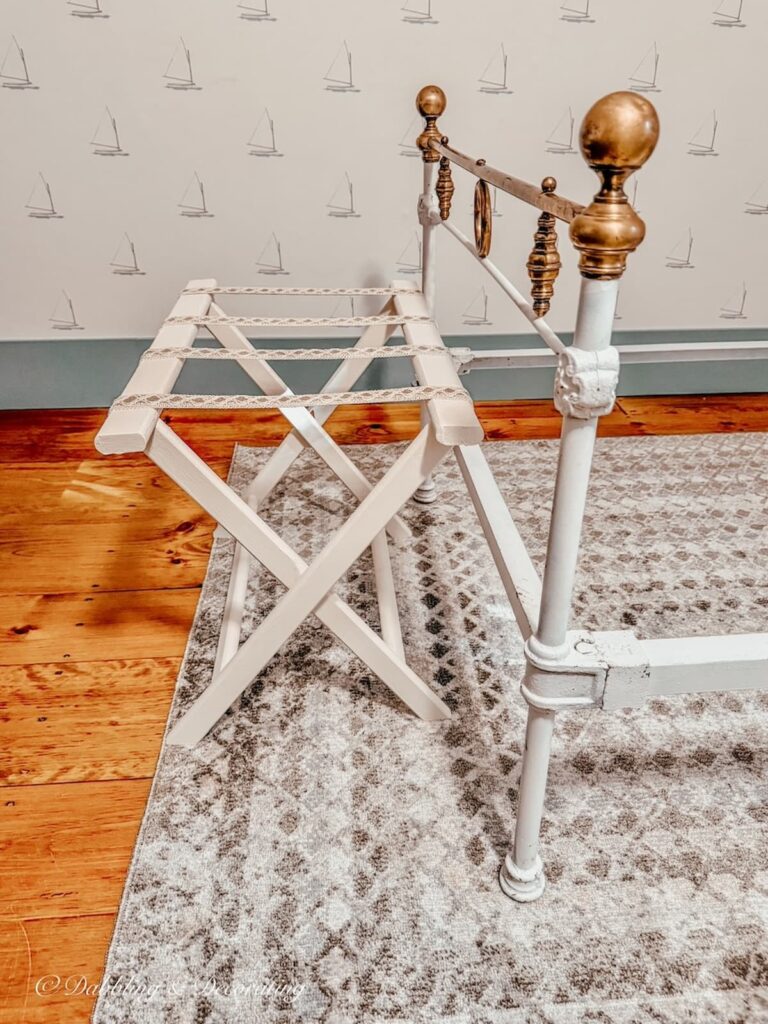 Folding suitcase holder painted white with ribbon at the foot of an antique white wrought iron bed with brass details in bedroom.